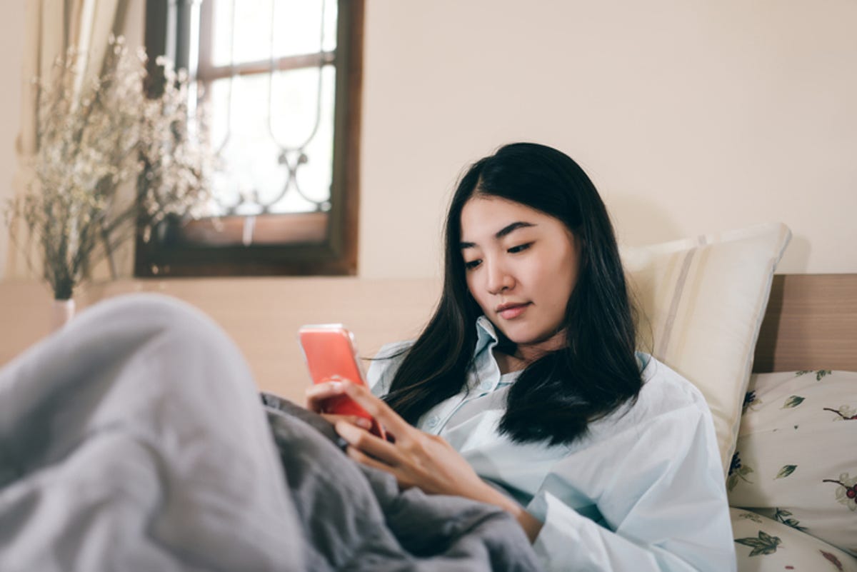 A young woman is sitting on the bed, looking at her phone.